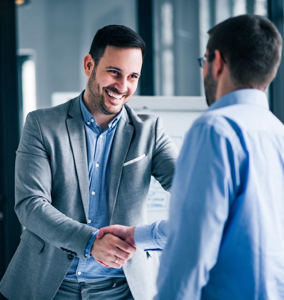two-smiling-businessmen-shaking-hands-while-standing-office (1) two-smiling-businessmen-shaking-hands-while-standing-office (1)
